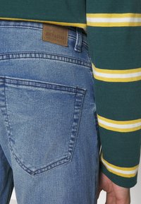 Blå denimjeans med en strukturert overflate, med et lærmerke merket "ONLY & SONS" på baksiden, kombinert med en grønnstripet langermet skjorte.