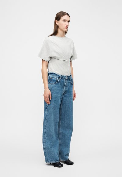 Γυναίκα όρθια, φοράει ανοιχτό γκρι κοντομάνικη μπλούζα με σκωληκοειδές σχέδιο και φαρδιά μπλε τζιν παντελόνια με μαύρα παπούτσια, σε λευκό φόντο.