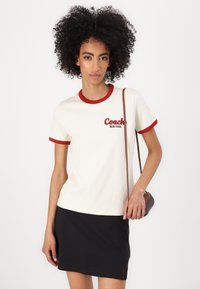 Kremfarget bomull T-skjorte med røde detaljer, med "Coach New York" broderi. Brukt med et svart miniskjørt og en liten brun skulderveske.