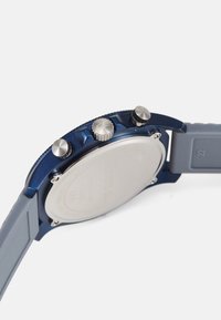 Reloj de metal azul con fondo plateado, presenta tres botones de presión, corona texturizada y correa de goma gris.
