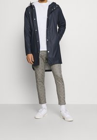 Marinblå vattentät jacka med huva, tryckknappar och medellång design; kombinerad med grårutiga byxor och vita sneakers.