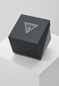 Juodas tekstūruotas dežutė su trikampiu dangčiu, ant viršaus esantis sidabrinis "GUESS" logotipas ir klausimo ženklo simbolis, pastatytas ant baltos ekspozicijos.