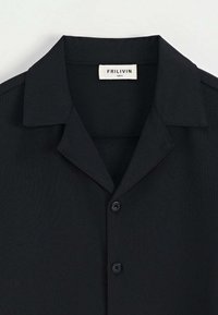 Chemise noire à manches courtes avec une texture douce, un col pointu et deux boutons noirs. L'étiquette indique la marque "FRILIVIN" et la taille "S".