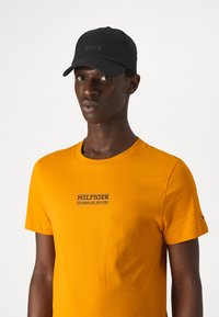 Cappello nero con logo "BOSS" e t-shirt di cotone arancione con grafica bianca e blu "HILFIGER" sul petto, maniche corte e scollatura rotonda.