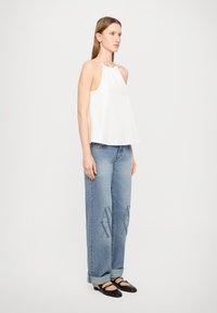 Weißes ärmelloses Oberteil mit hohem Ausschnitt, kombiniert mit hellblauen weiten Jeans, die ein ausgewaschenes Muster und hochgekrempelte Säume aufweisen. Schwarze flache Schuhe.