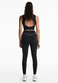 Black sports bra with open back and white stitching, paired with black leggings. Both made from a stretchy material, accentuating form.