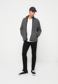 Hombre con una gorra oscura, chaqueta de forro polar gris con cremallera, camiseta blanca, pantalones ajustados negros y zapatillas negras, de pie con las manos en los bolsillos.
