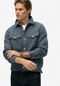 Szary, teksturowany podkoszulek z dwoma przednimi kieszeniami, kołnierzykiem zapinanym na guziki i długimi rękawami, noszony na białej koszuli i zestawiony z ciemnymi dżinsami.