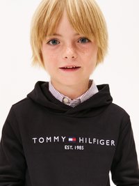 Melnā kapuce ar izšūtu tekstu "TOMMY HILFIGER EST. 1985" baltā krāsā, uzvilkta virs svītrainas krekla ar apkakli. Gaišblondīni mati un zilas acis.