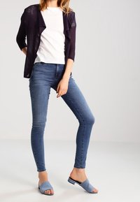 Cardigan blu navy indossato sopra una T-shirt bianca, abbinato a jeans aderenti blu e sandali slip-on azzurri. Design semplice e lineare.