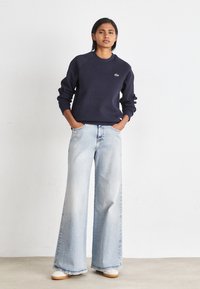 Marineblå hettegenser med rund hals og logo, kombinert med lyseblå vide bukser og beige sneakers. Glatt tekstur, avslappet stil.