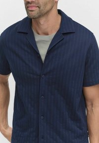 Marineblauw overhemd met korte mouwen, voorzien van een gestructureerd streepjespatroon, met een knoopsluiting aan de voorkant en een nonchalante kraag. Gemaakt over een lichtgrijs shirt.