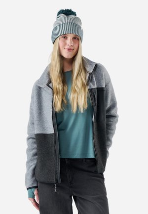 Joven mujer con pelo rubio largo que lleva una chaqueta con cremallera gris y negra, camiseta color azul verdoso, pantalones negros y un gorro de punto a rayas con pompón.
