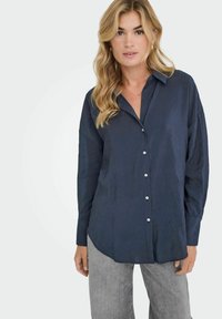 Donna bionda con una camicia a maniche lunghe blu navy sbottonata e pantaloni grigio chiaro, in posa contro uno sfondo bianco semplice.