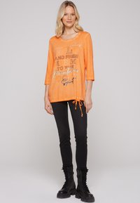 Leichtes oranges Oberteil mit rundem Halsausschnitt, Dreiviertelärmeln und Kordelzugsaum. Versehen mit bedrucktem Text in kontrastierenden Farben.