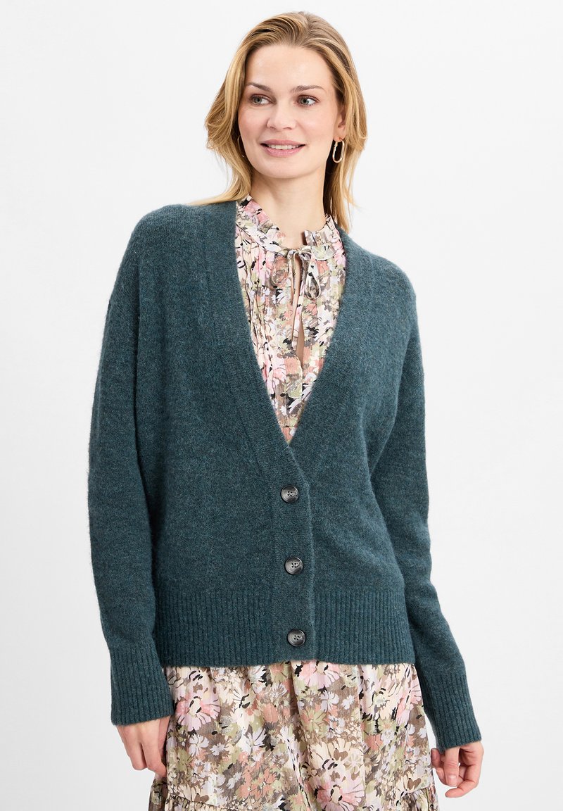 Teal wollen cardigan met een V-hals, voorzien van vier donkere knopen en geribde manchetten. Gekocht over een jurk met een bloemenprint in roze tinten.