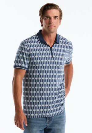 Mann trägt ein kurzärmeliges, blaues Poloshirt mit geometrischem Muster, Kragen und Reißverschluss, steht mit einer Hand in der Tasche vor weißem Hintergrund.