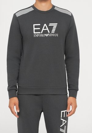 Dunkelgraues Sweatshirt und passende Hose mit weißen EA7 Emporio Armani-Logos auf Brust und Oberschenkel, getragen von einer stehenden Person.