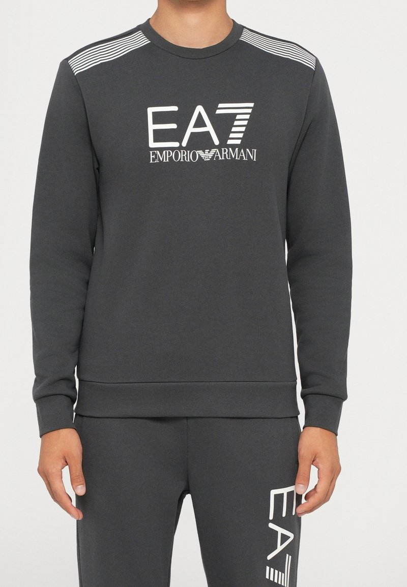 Sweat-shirt gris foncé et pantalon assorti avec logos blancs EA7 Emporio Armani sur la poitrine et la cuisse, portés par une personne debout.