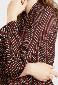 Röd och vit mönstrad blus med volangkrage och långa ärmar, som har en zickzackdesign och mjuk, flödande tyg.