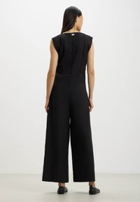 Tuta nera con un corpetto aderente e pantaloni a gamba larga. Presenta maniche a cappuccio e una cerniera sul retro. Tessuto liscio con dettagli minimi.