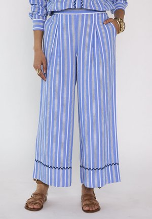 Blauw-witte, verticaal gestreepte broek met wijde pijpen en zwarte golvende rand aan de zoom, gedragen met bruine sandalen en gouden armbanden en ringen.