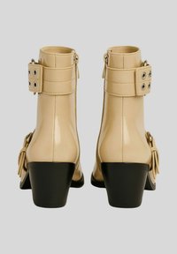 Stivali alla caviglia in pelle verniciata beige con tacco medio, presentano un dettaglio con fibbia, cerniere laterali e una suola nera a contrasto.