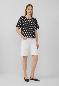 Schwarzes Oberteil mit weißen Punkten, kurzen Ärmeln und einem Schlüsselloch-Ausschnitt. Kombiniert mit weißen, hochgerollten Shorts und schwarzen spitzen Ballerinas.