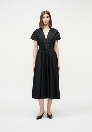 Femme debout portant une robe midi noire avec un décolleté en V profond, une taille cintrée par des boucles, et des manches courtes, associée à des escarpins noirs à bout pointu.