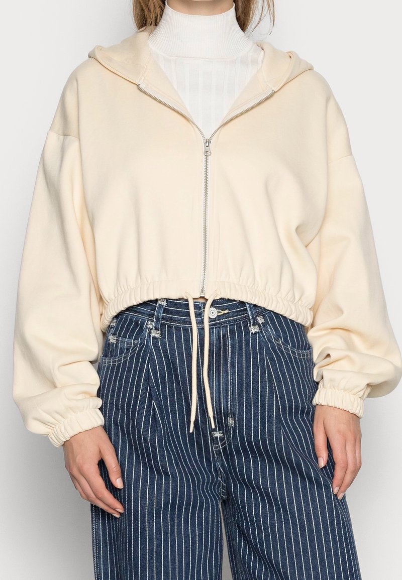 Žena v krémové zkrácené mikině na zip přes bílé žebrované rolákové tričko, doplněná vysokými tmavě modrými proužkovanými džíny.