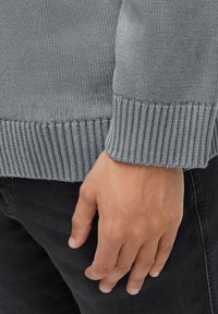 Grauer gestrickter Pullover mit geripptem Saum, im Nahaufnahme eine Hand, die auf schwarzen Hosen ruht. Die Textur wirkt weich und atmungsaktiv.