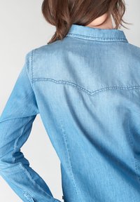 Chemise en denim bleu clair avec un col pointu, des poignets boutonnés et un design de couture sur le dos. Texture lisse avec un effet de dégradé subtil.