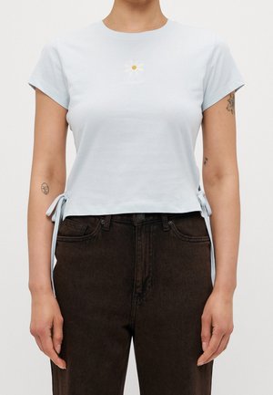 Jasnoniebieski krótki t-shirt wykonany z bawełny, z haftem stokrotki i wiązaniami po bokach, w połączeniu z ciemnoskórzanymi dżinsami.