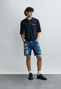 Crna polo majica s kratkim rukavima i bijelim logotipom, uparena s plavim traper kratkim hlačama s višebojnim tiskanim detaljima i crnim tenisicama.