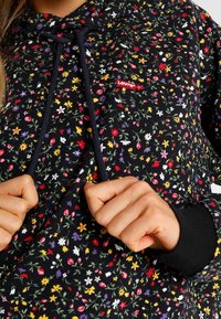 Mains tenant les cordons d'une capuche noire sur un sweat-shirt Levi's noir à imprimé floral avec un patch logo rouge sur la poitrine.
