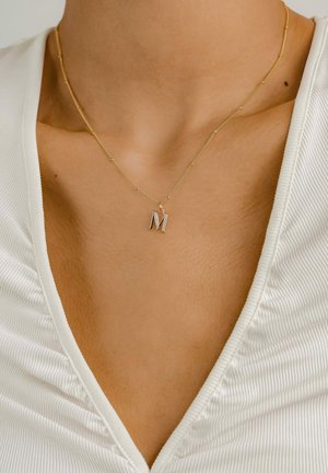 Collier en or avec un pendentif en forme de "M" cursif orné de diamants. L'arrière-plan est un haut texturé de couleur claire avec un décolleté en V.