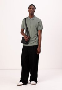 Olivenfarget polo-skjorte med glidelås-krage, kombinert med løse svarte bukser. Svart crossover-veske og hvite sneakers med beige detaljer.