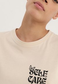 Beige t-shirt featuring a round neckline, short sleeves, and a black “SELF CARE” graphic with a butterfly detail on the chest area.