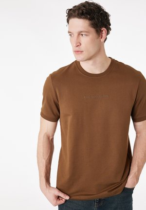 Giovane uomo con capelli ricci che indossa una t-shirt marrone a maniche corte con la scritta "ECHO THROUGH TIME", guarda verso sinistra, tenendo l'orlo della maglietta con la mano.