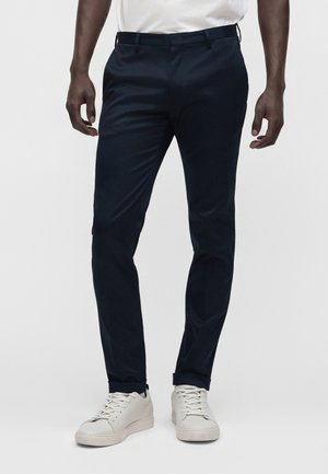 Homme portant un pantalon slim bleu foncé et des baskets blanches, debout devant un fond clair uni.