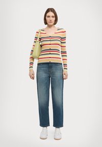 Gestreepte, geribbelde lange mouwen cardigan in geel, rood, zwart en groen, gecombineerd met lichtblauwe rechte jeans en witte sneakers. Licht schoudertas.