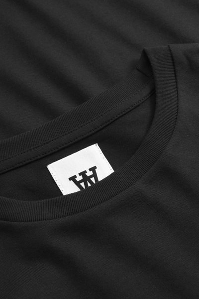 Camiseta negra de algodón con cuello redondo acanalado y una etiqueta blanca con letras negras. Textura suave sin patrones visibles.