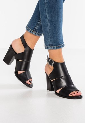 Personne portant un jean bleu et des sandales à talons bloc ouvertes noires avec des brides à la cheville, se tenant sur un sol blanc.