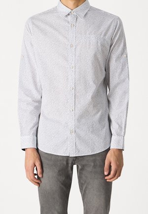 Uomo che indossa una camicia bianca a maniche lunghe con piccoli puntini neri e jeans grigi, in piedi con le braccia rilassate.