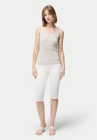 Ung kvinna klädd i ärmlös beige ribbad topp, vita knälånga smala byxor och beiga öppna sandaletter med blockklack, stående mot en enkel bakgrund.