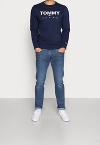 Marinblå sweatshirt med vit "TOMMY JEANS"-logga, i kombination med ljusblå denimjeans och vita sneakers. Enkel, avslappnad passform och design.