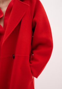 Cappotto rosso di lana con un colletto appuntito, un unico bottone nero e una vestibilità comoda. Tessuto morbido e maniche di lunghezza intermedia.