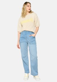 Maglione lavorato a maglia crema con la scritta "Estate" in giallo, abbinato a jeans a gamba larga azzurri chiari con tasche frontali e cuciture a vista.