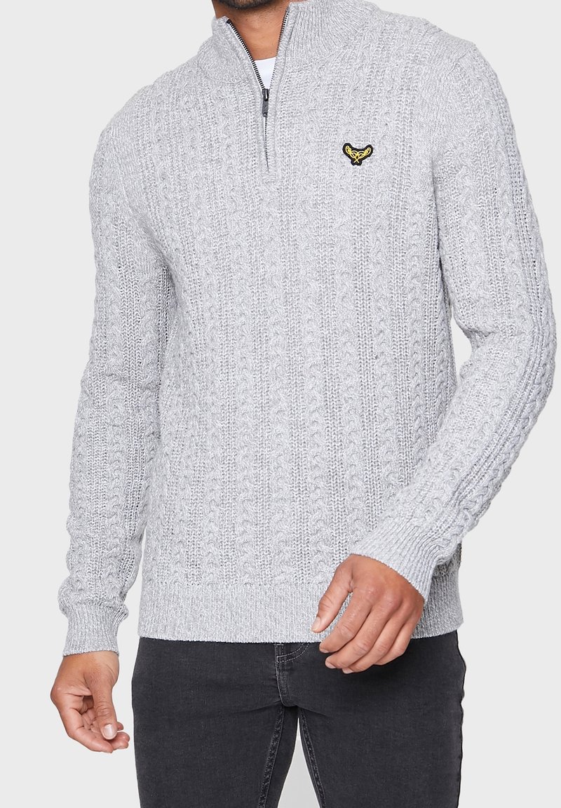 Homme portant un pull gris clair en maille torsadée avec une fermeture éclair courte et un petit logo brodé sur la poitrine, associé à un jean foncé.