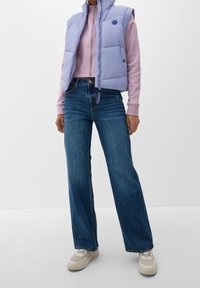 Gilet imbottito di un colore viola chiaro sopra una felpa rosa con zip, abbinato a jeans a gamba larga blu e sneakers color crema.